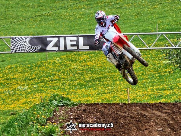 Motorsport Event mit Sprung: Ein Fahrer fliegt vor dem Ziel-Banner über die blühende Löwenzahnwiese