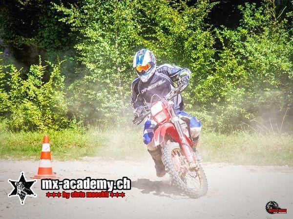 Motocross Fahrer in blauer Jacke an einer Pylone, aufgewirbelter Staub vor grünen Büschen