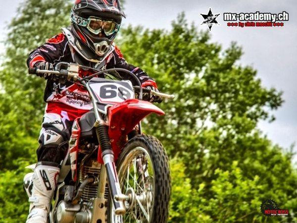 Jugendlicher Fahrer mit Startnummer 68 auf einem roten Motocross-Motorrad vor einem grünen Baum
